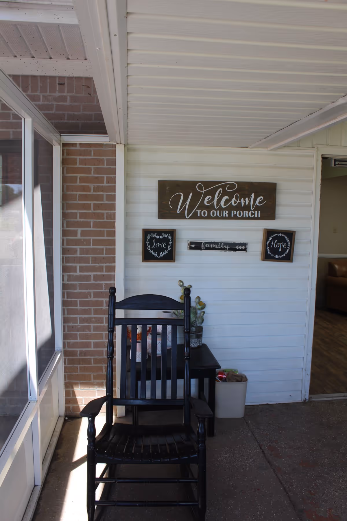 Black rocking chair on a covered porch in front of a wall with a 'Welcome to Our Porch' sign and a small side table.