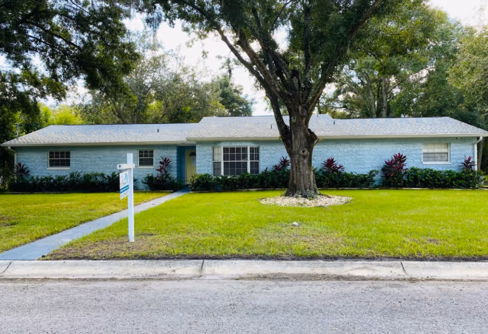 Single-story light blue brick building with a gray roof, surrounded by green grass and trees. A large tree is in the front yard with a small white rock border around its base. A concrete walkway leads from the sidewalk to the front door. There is a white signpost near the sidewalk in front of the building.