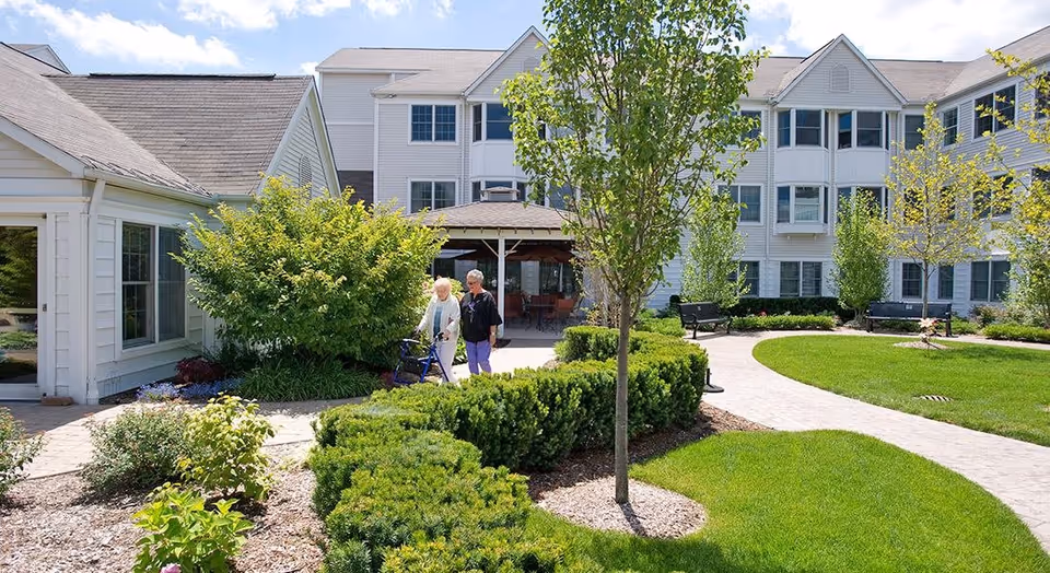 Outdoor garden area at The Orchards at Canterbury-on-the-Lake with a paved walkway, green grass, bushes, and trees. Two elderly women are walking along the path near a white building with multiple windows and a covered seating area.