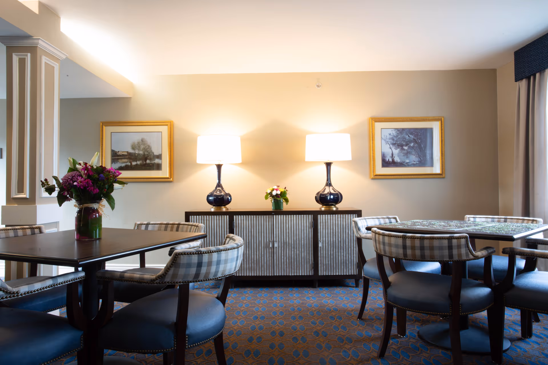 A well-lit interior room with two tables surrounded by cushioned chairs with plaid backs. On one table is a vase with purple flowers. Against the beige wall is a sideboard with two matching lamps and a small flower arrangement in the center. Two framed landscape paintings hang on the wall above the sideboard. The floor is carpeted with a patterned design.