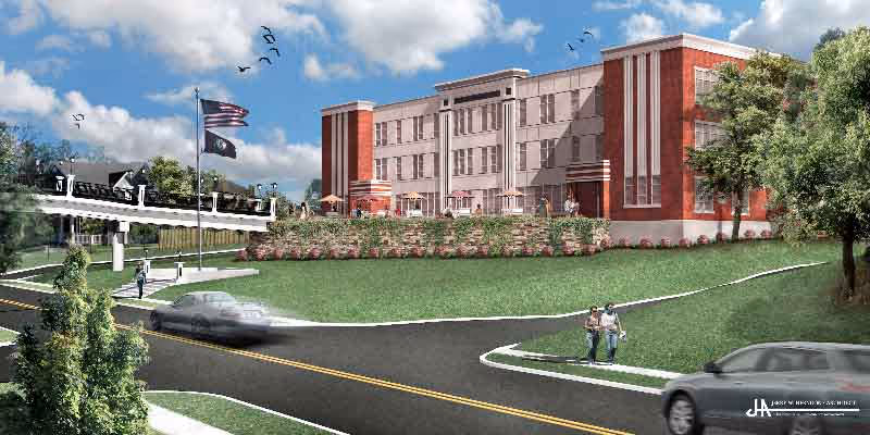 Exterior view of a large, multi-story brick building with a manicured lawn and landscaping in front. Two flags are flying on flagpoles near the building, and a few people are walking on the sidewalk. Cars are driving on the road in front of the building under a partly cloudy sky.