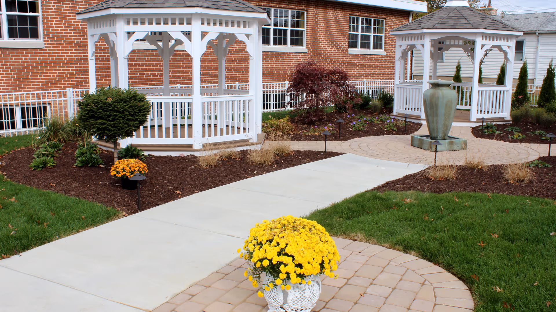 Outdoor garden area at Palmyra Heritage House featuring two white wooden gazebos, a large green ceramic urn fountain, paved walkways, landscaped flower beds with shrubs and yellow flowers, and a brick building in the background.