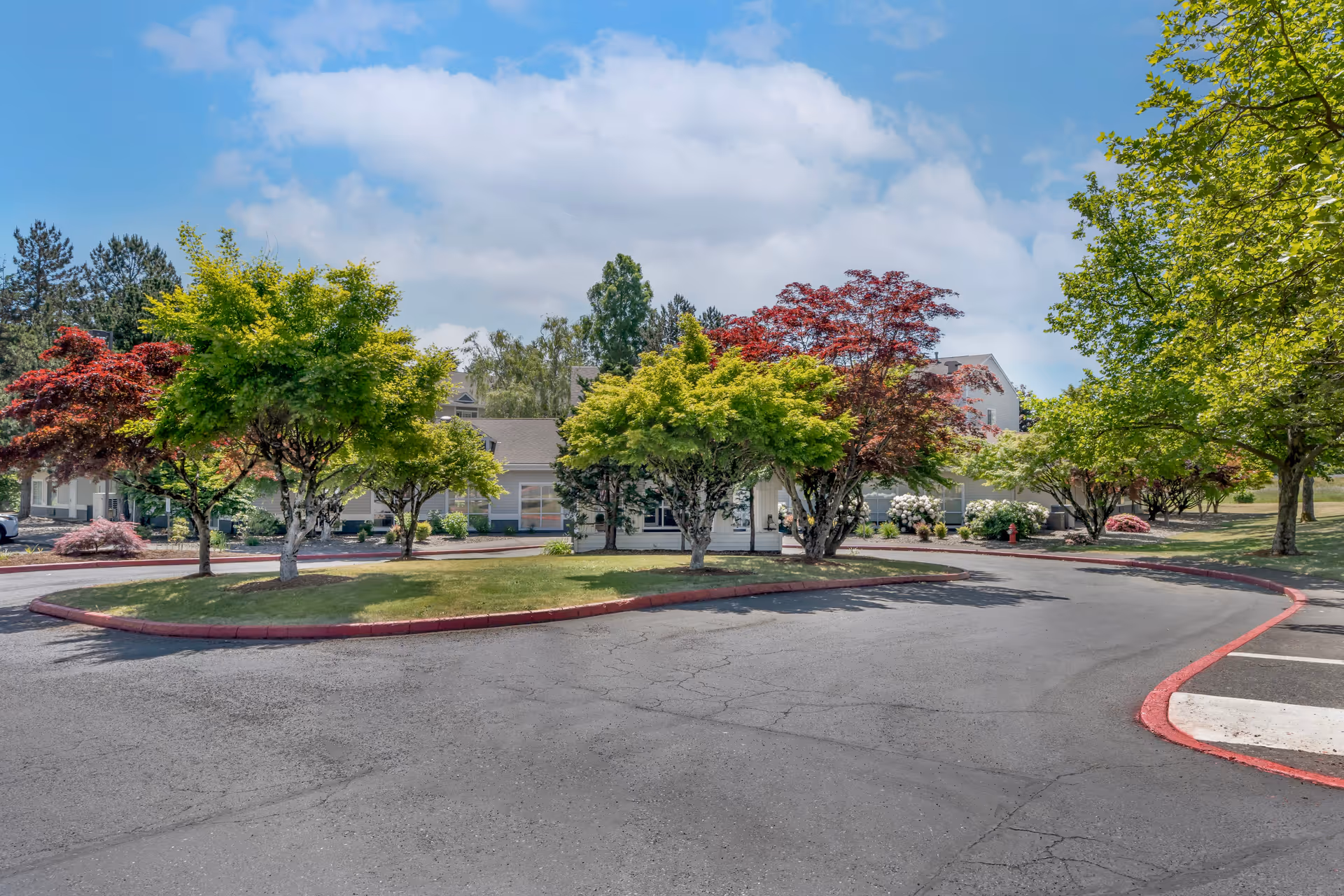 Exterior view of Brookdale Mt. Hood facility showing a driveway with landscaped islands featuring green and red-leaved trees, a parking area, and a partly cloudy sky.