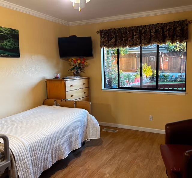 Assisted living bedroom with a single bed, wooden dresser topped with a TV and flowers, a window showing an outdoor garden, and a chair.