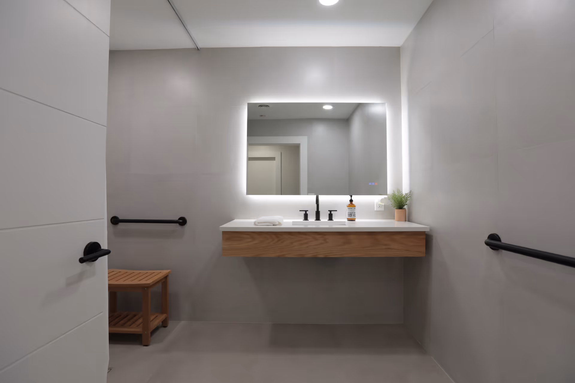 A modern bathroom with light gray tiled walls and floor, featuring a floating wooden vanity with a white countertop. On the countertop, there is a folded white towel, a soap dispenser, and a small potted plant. Above the vanity is a large rectangular mirror with backlighting. There are black grab bars on both side walls and a wooden bench in the corner. A white door with a black handle is partially open on the left side.
