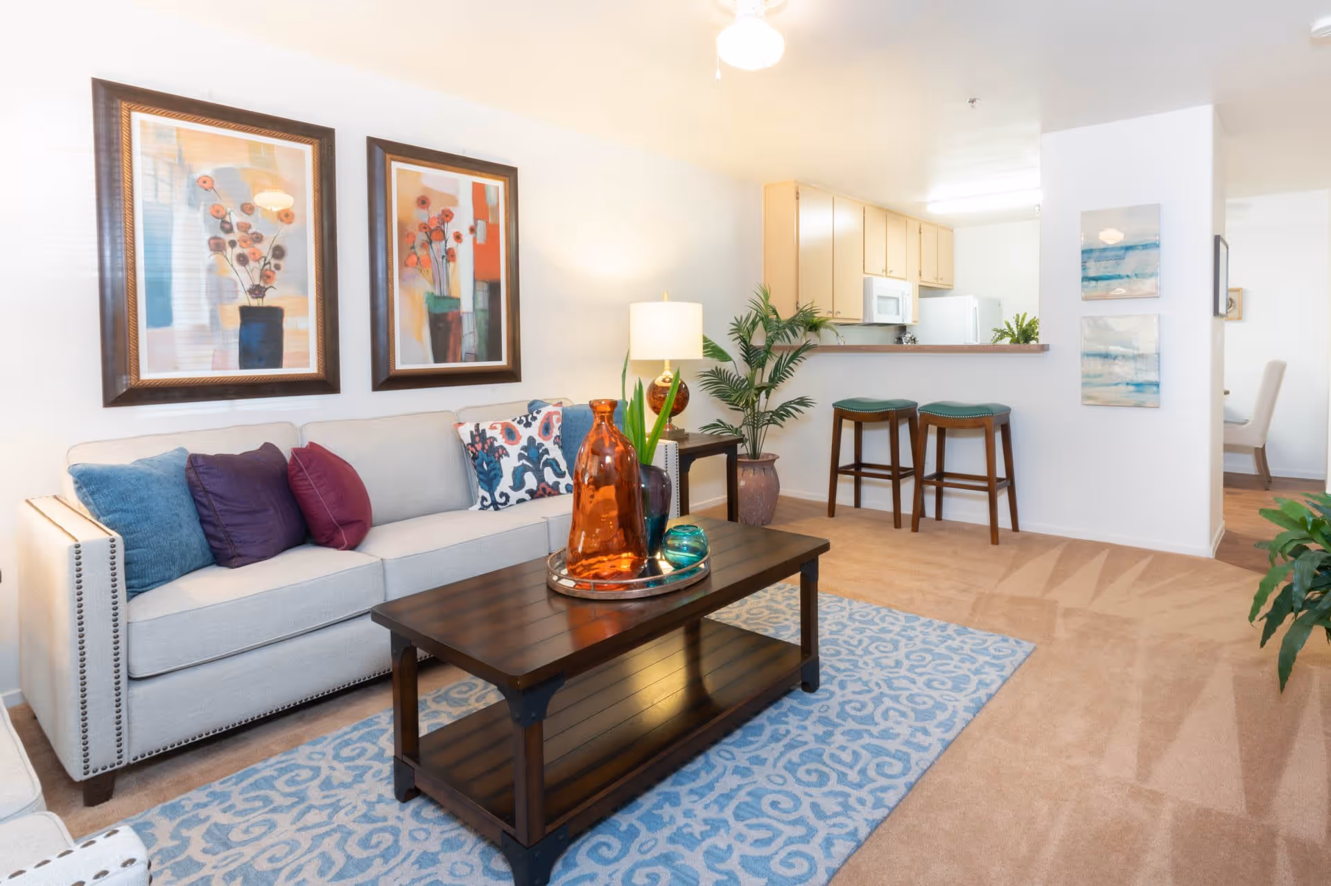 A bright and cozy living room featuring a light gray sofa with colorful cushions, a dark wooden coffee table with decorative glass vases, a blue patterned rug, and two framed floral paintings on the wall. In the background, there is a kitchen area with a counter, two bar stools, and light wood cabinetry. The room is carpeted and decorated with green plants and soft lighting.