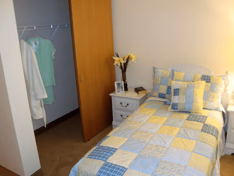 A small bedroom with a single bed covered in a patchwork quilt with yellow, blue, and white squares. There are matching pillows on the bed. Next to the bed is a white nightstand with two drawers, a framed photo, a book, and a vase with yellow flowers. An open closet with a wooden sliding door is visible, containing a hanging green outfit and a white robe.
