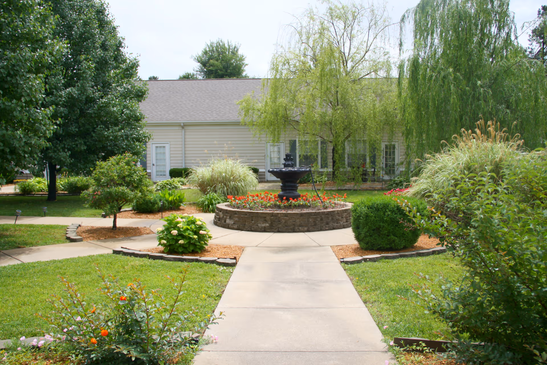 Landscaped outdoor courtyard with a central fountain, flowerbeds, and a walkway leading to a single-story building.