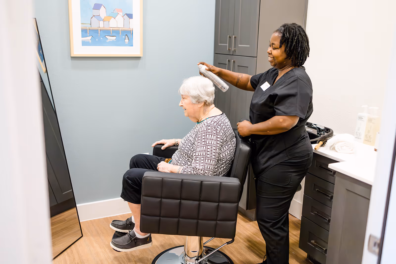 A senior woman with white hair sits in a black salon chair while a smiling hairstylist sprays her hair in a small, well-lit room with light blue walls, a large mirror, and a framed picture of houses by the water.