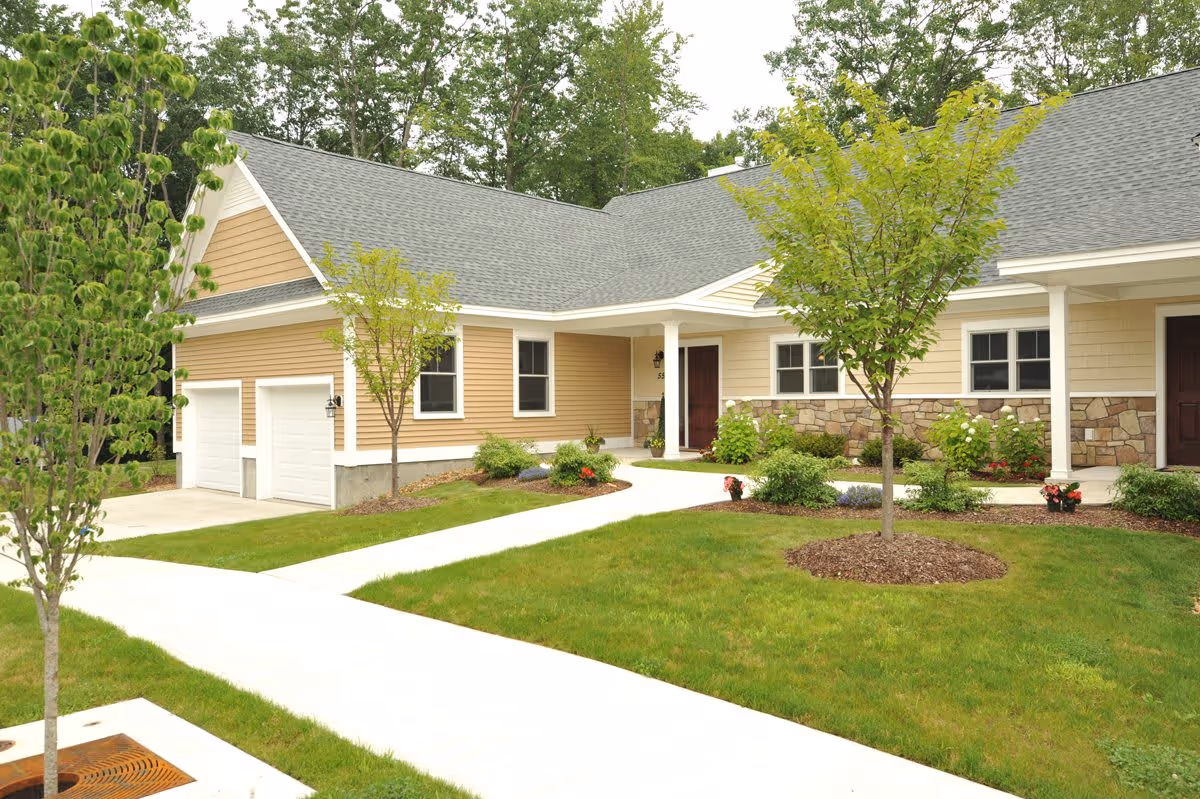 Exterior view of a single-story residential building with beige siding, stone accents, and a gray shingled roof. The building has two white garage doors, a covered entrance with a dark brown door, and landscaped green lawns with small trees and shrubs. A concrete walkway leads to the entrance.