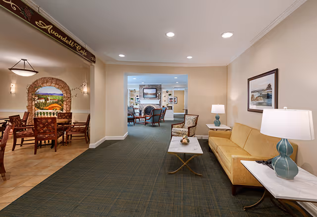 Interior view of a senior living facility showing a hallway with a seating area on the right featuring a yellow sofa, two side tables with lamps, and a framed picture on the wall. On the left, there is an entrance to the Avondell Cafe with a dining area that has wooden chairs and tables, and a mural on the wall. Further in the background, there is another common area with chairs and a fireplace.
