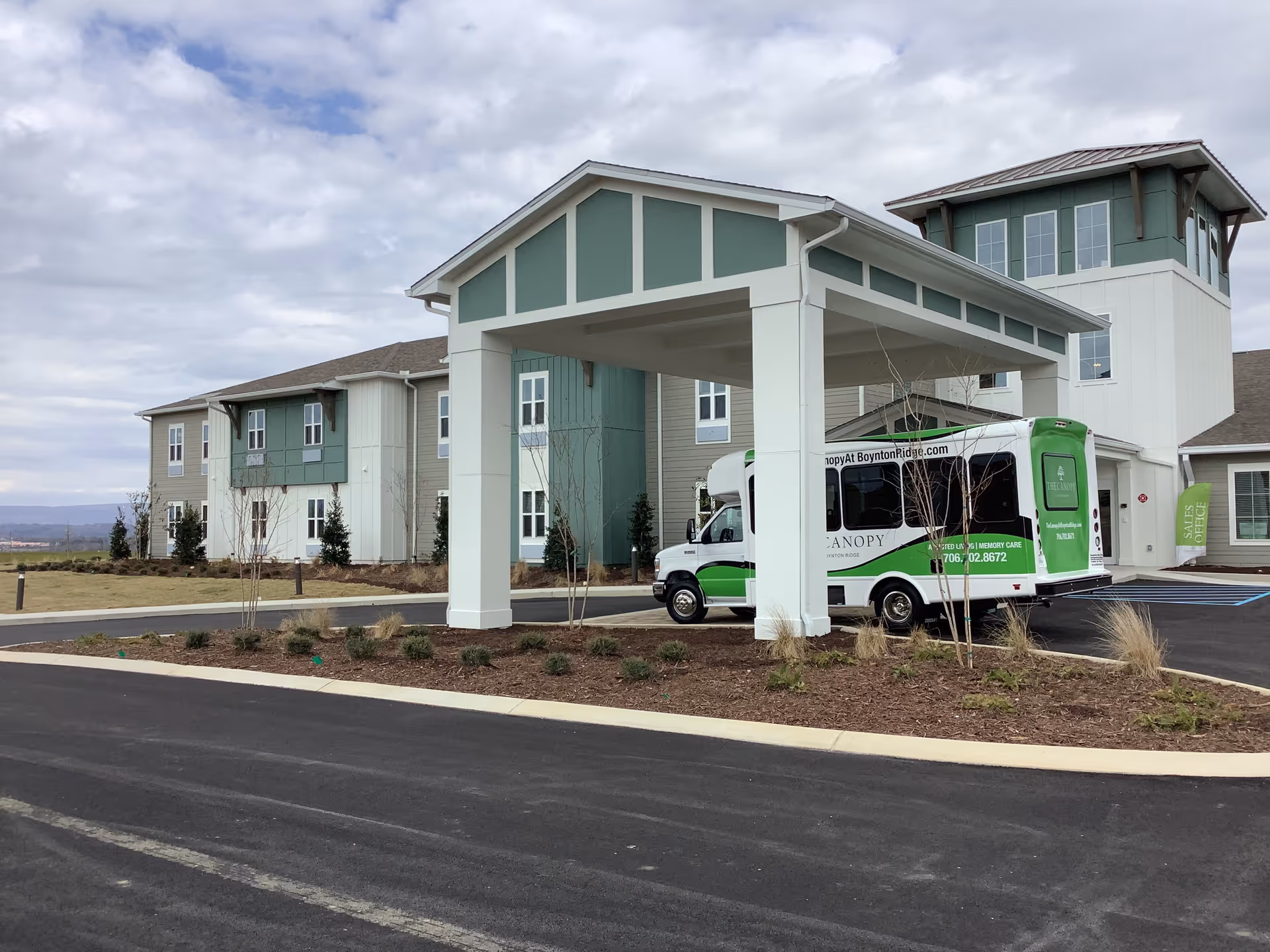 Front entrance of a senior living building with a covered canopy and a green-and-white shuttle bus parked underneath.