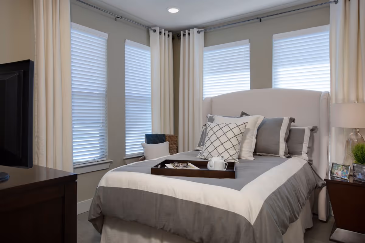A neatly made bed with gray and white bedding and multiple pillows in a bedroom with three windows covered by white blinds and cream-colored curtains. There is a wooden nightstand with a lamp, framed photos, and a small plant on the right side of the bed, and a dark wooden dresser with a TV on the left side.