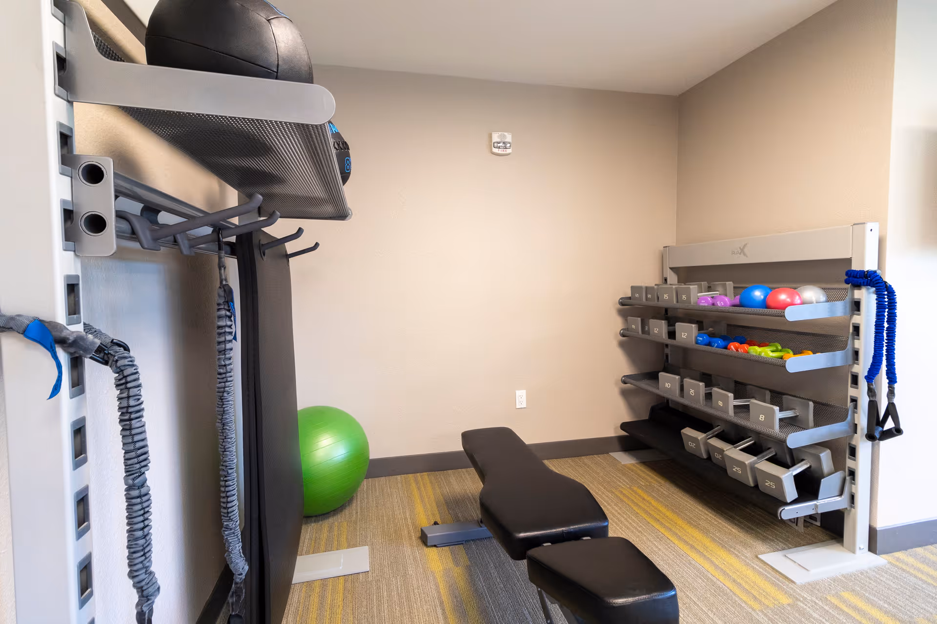 A small fitness room with a black workout bench in the center, a green exercise ball against the wall, resistance bands hanging on the left wall, and a rack on the right holding various dumbbells and colorful exercise balls.