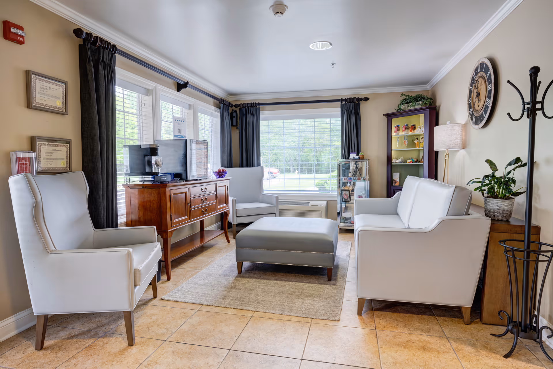 A bright and cozy living room with large windows covered by dark curtains. The room features two white armchairs, a white loveseat, and a gray ottoman on a beige rug. There is a wooden sideboard with a coffee machine and other items, a tall glass display cabinet, a purple shelving unit with decorative items, a floor lamp, a wall clock, and a coat rack. The floor is tiled in a light brown color.
