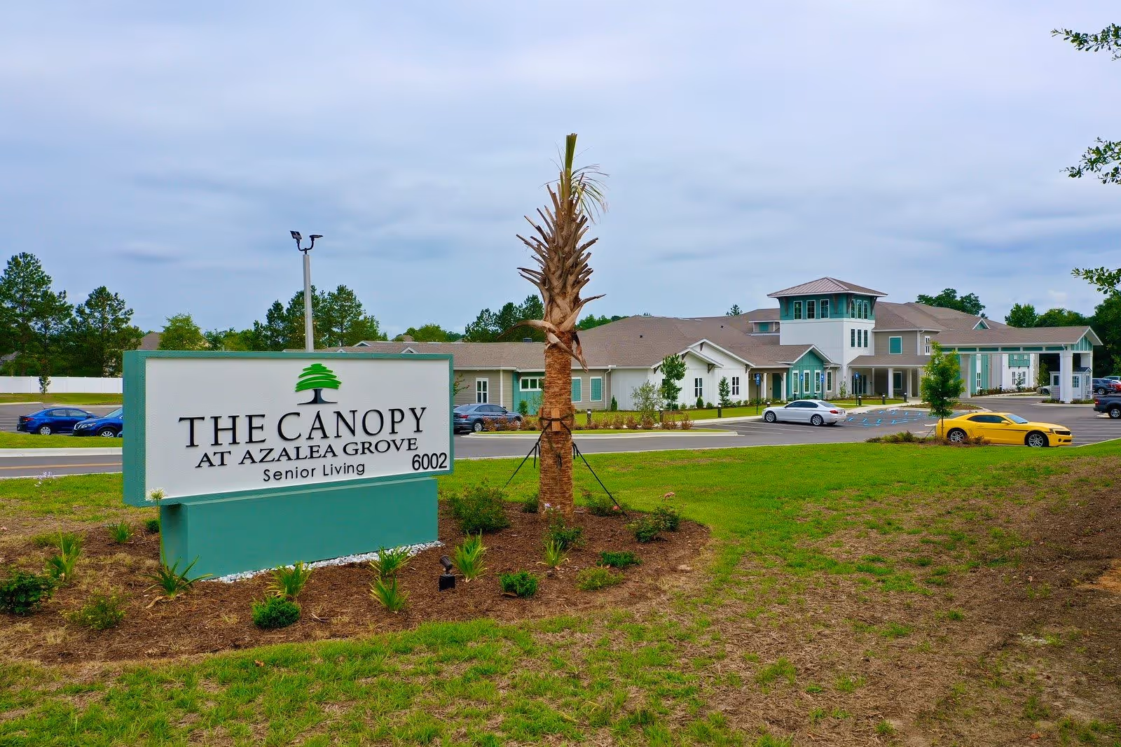 Exterior view of The Canopy at Azalea Grove senior living facility with a large sign in the foreground displaying the facility's name and address. The building is surrounded by a parking lot with several cars and landscaped greenery under a cloudy sky.