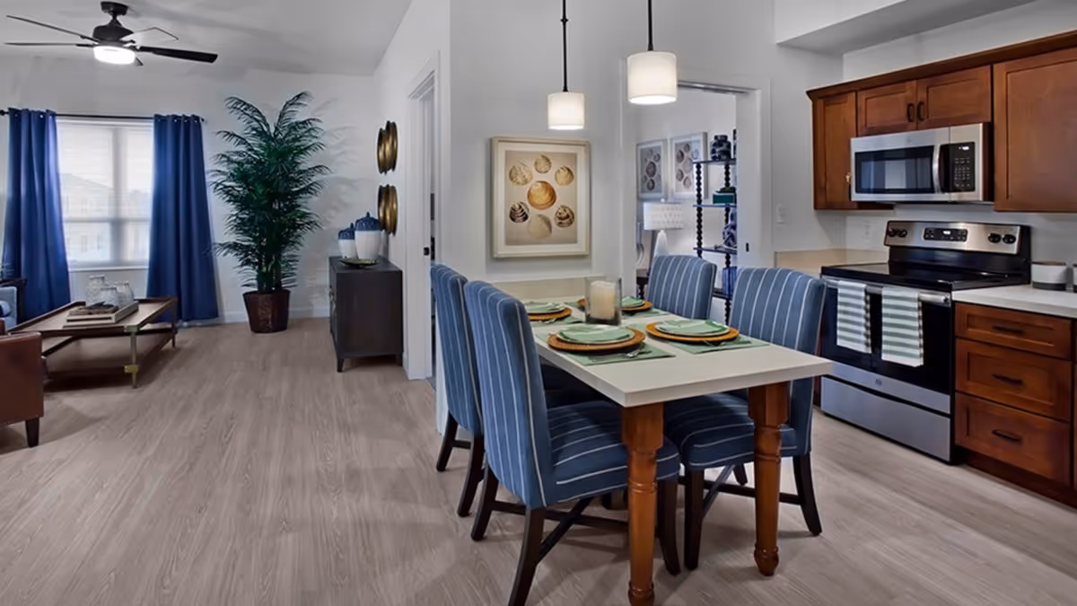Interior view of a senior living facility showing a dining area with a table set for four with blue striped chairs, adjacent to a kitchen with wooden cabinets, a stainless steel stove, and microwave. In the background, there is a living area with a ceiling fan, blue curtains, a large potted plant, and a small cabinet with decorative items.