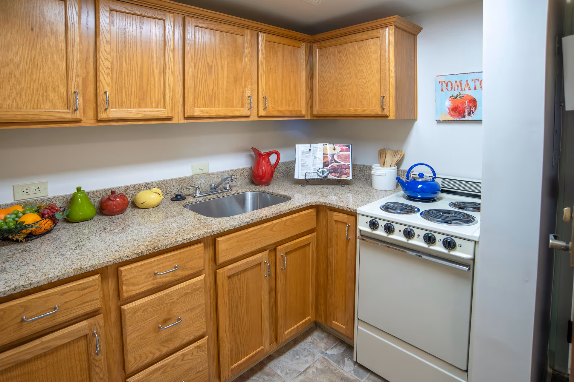 A small kitchen with wooden cabinets, a granite countertop, a stainless steel sink, an electric stove with a blue kettle on it, and various decorative items including a red pitcher, ceramic fruit, and a recipe book on a stand. There is a painting of a tomato on the wall.
