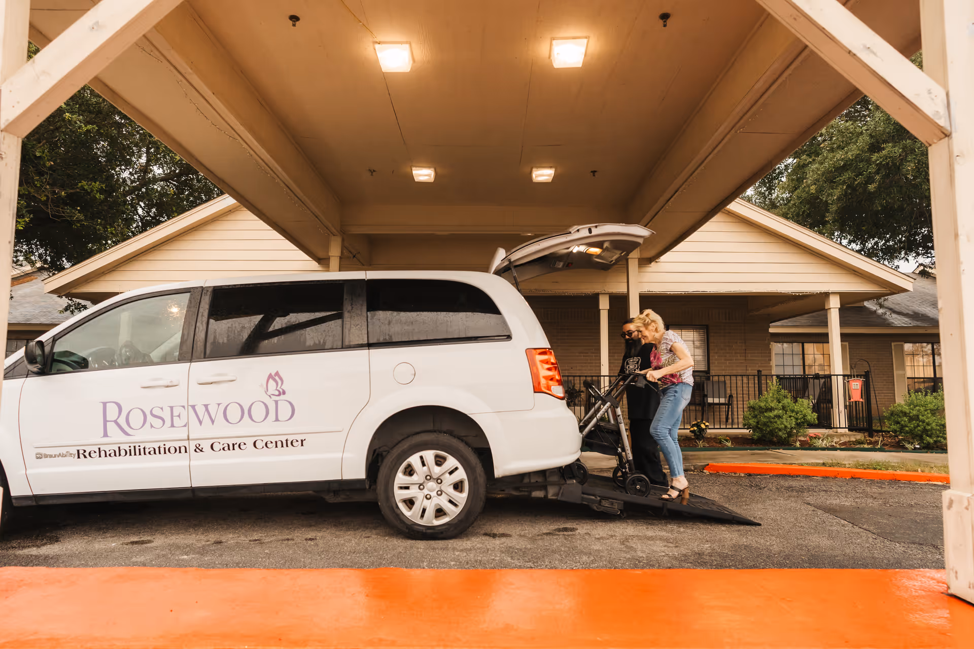 A white van with the logo and text 'Rosewood Rehabilitation & Care Center' is parked under a covered drop-off area. Two women are assisting a person using a wheelchair ramp to enter the van. The background shows a building with beige siding and some greenery.