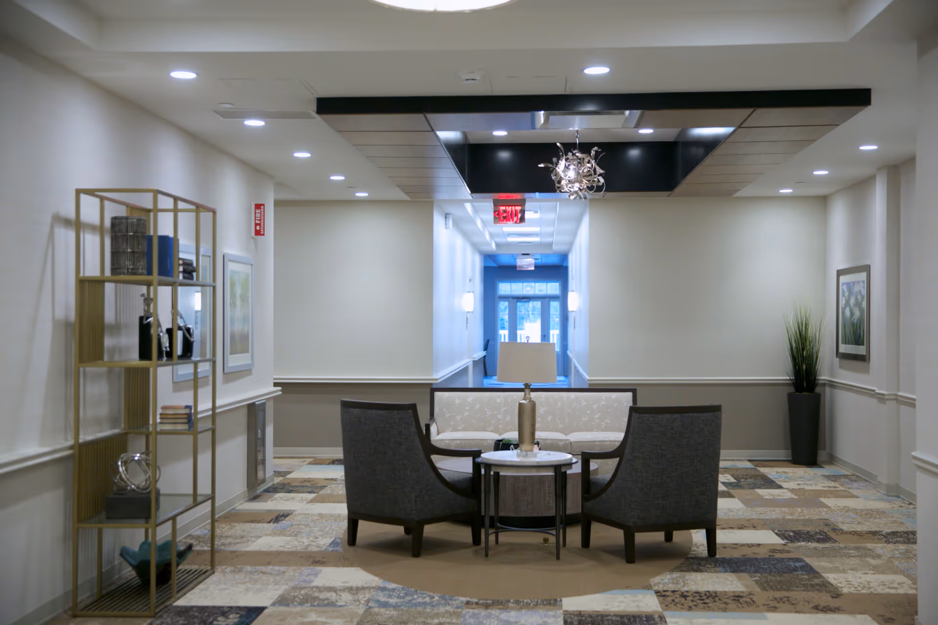 A well-lit interior hallway seating area with two gray armchairs and a white patterned sofa arranged around a small round table with a lamp. The floor has a patterned carpet, and there is a decorative shelving unit with books and ornaments on the left side. The hallway extends to a door with windows at the far end, and there is a modern chandelier hanging from the ceiling.