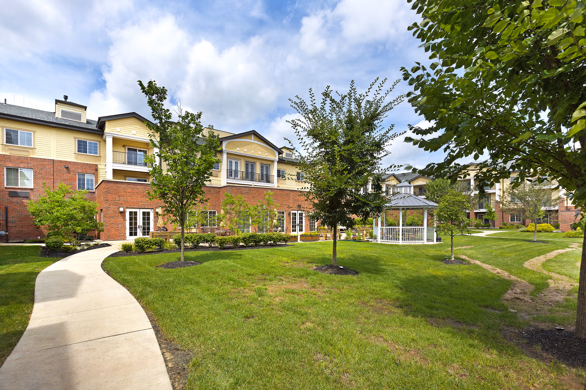 Outdoor view of a senior living facility with a well-maintained lawn, several young trees, a white gazebo, and a paved walkway leading through the garden area. The building in the background has a brick lower level and light yellow upper levels with balconies and windows under a partly cloudy sky.