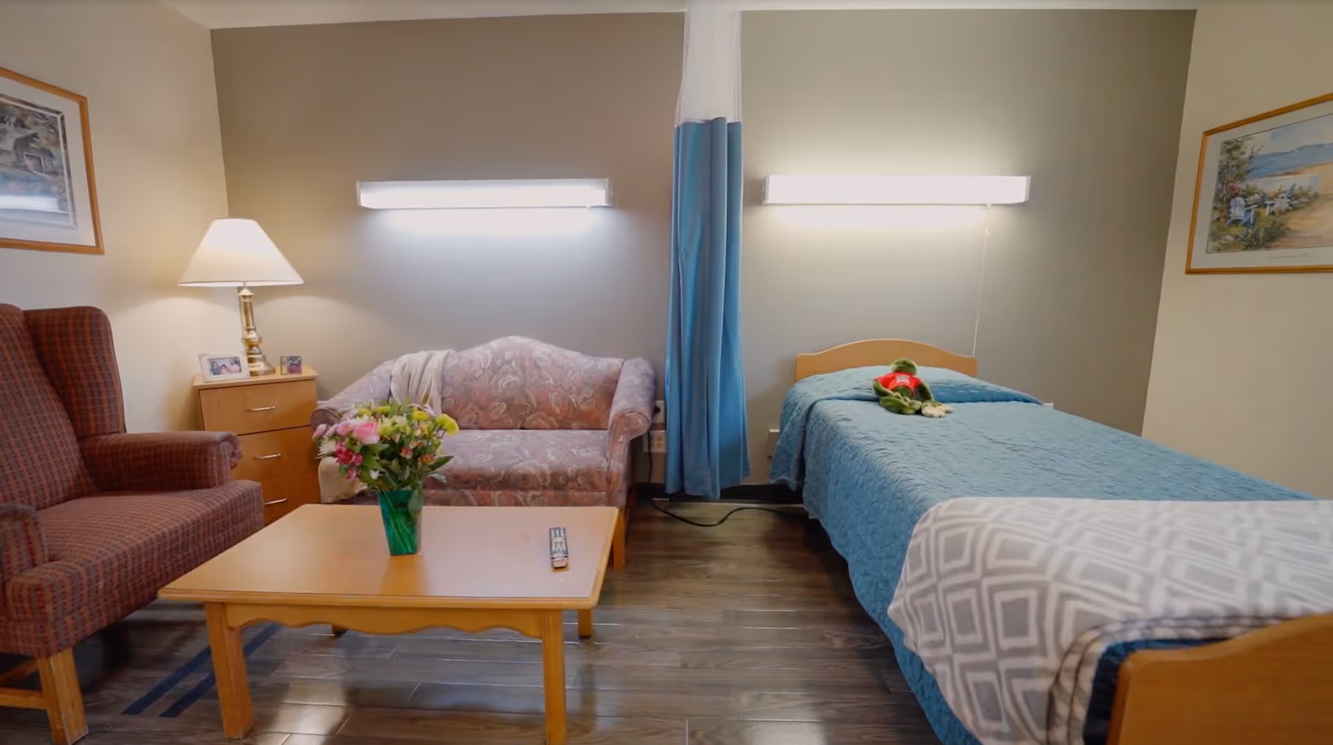 A cozy room with a single bed covered in a blue quilt and a gray patterned blanket, a small sofa with floral upholstery, a wooden coffee table with a vase of flowers and a TV remote, a red armchair, a wooden nightstand with a lamp and framed photos, and two wall-mounted lights above the bed and sofa. The room has wooden flooring and framed artwork on the walls.