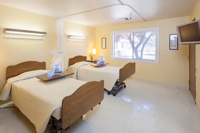 A bright room with two hospital-style beds with wooden headboards and footboards, each covered with beige quilts. Each bed has a small overbed table with a gift basket on top. The room has pale yellow walls, a large window with a view of trees outside, a wall-mounted TV, and two wall lights above the beds.