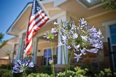 Front view of a senior living facility with an American flag and purple flowers.