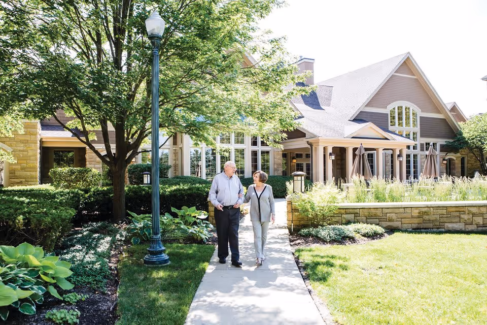 An elderly couple walking hand in hand on a paved pathway surrounded by green bushes and trees, with a large building featuring tall windows and a porch in the background under a sunny sky.