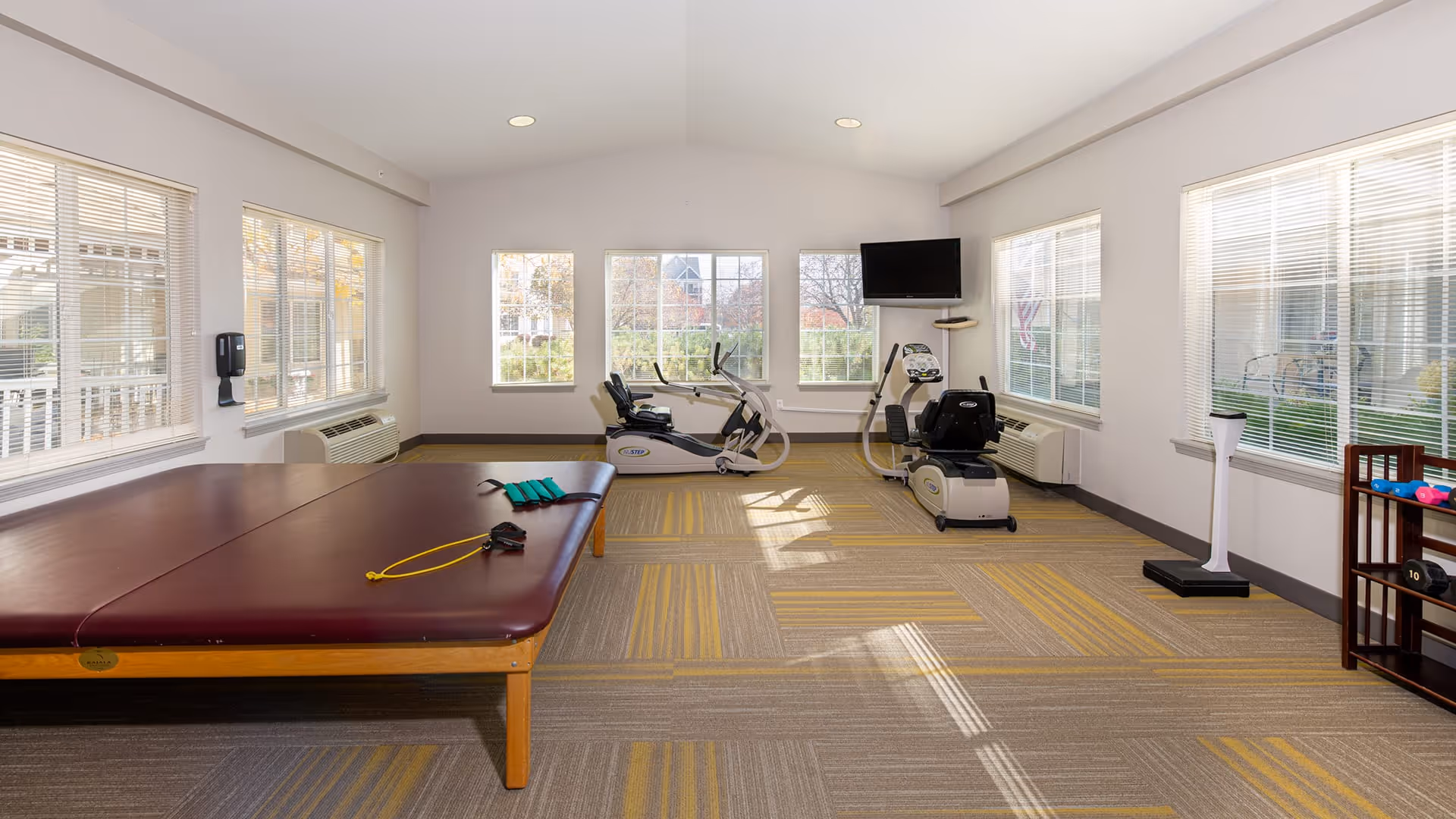 A bright exercise room with large windows on three walls letting in natural light. The room contains two recumbent exercise bikes, a large padded exercise table with resistance bands on it, a TV mounted on the wall, and a small rack holding various dumbbells and exercise equipment. The floor is carpeted with a pattern of gray and yellow squares.
