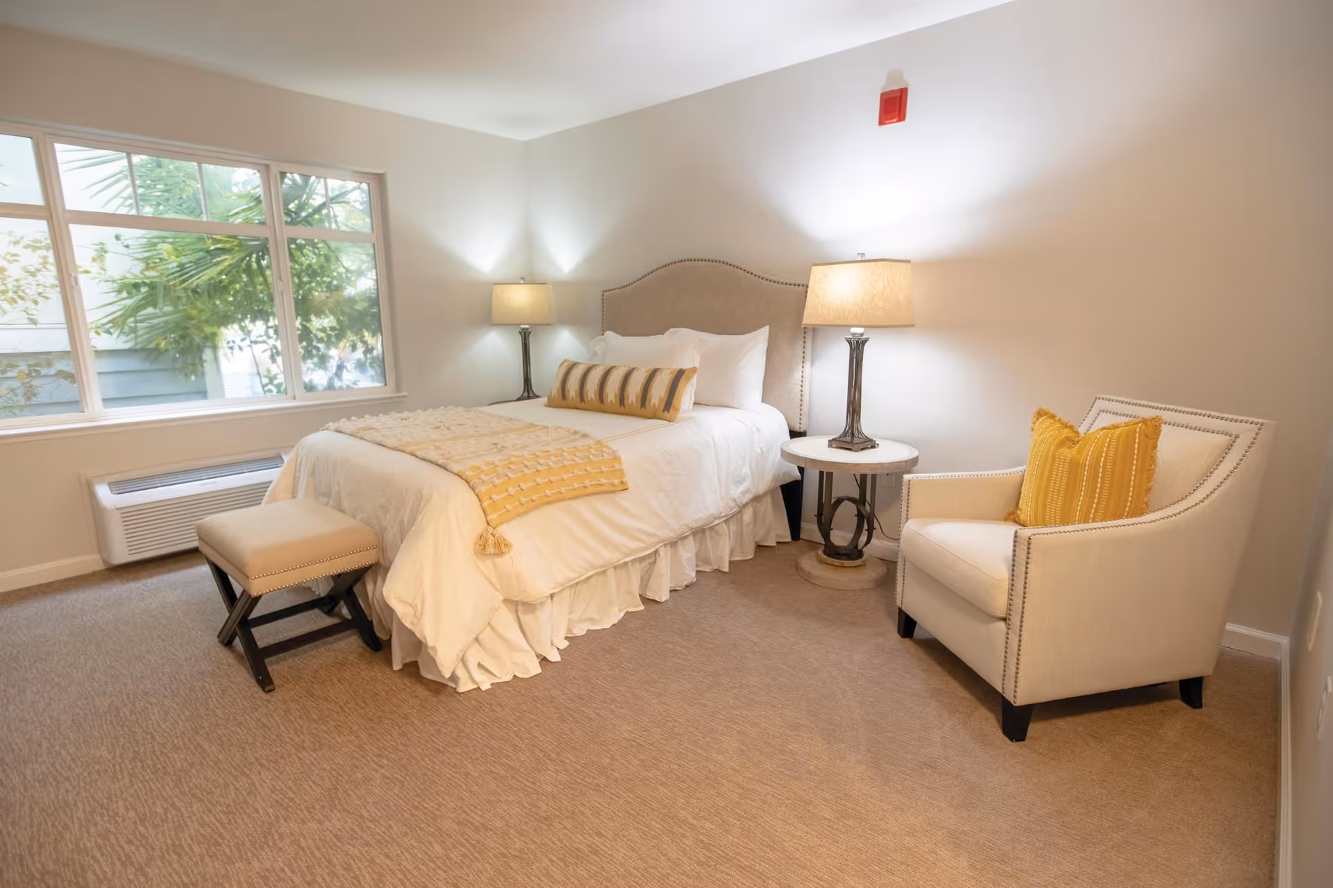 Bright furnished bedroom with a bed, two lamps on nightstands, an upholstered armchair with a yellow pillow, and a large window.