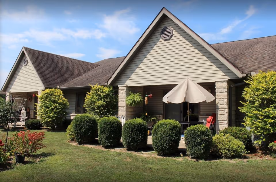 Exterior view of a single-story building with beige siding and a brown shingled roof. The building has a covered patio area with an umbrella, outdoor chairs, and various plants and bushes in front. The sky is partly cloudy and the lawn is green and well-maintained.