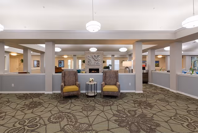 A spacious senior living facility common area with patterned carpet, two brown armchairs with yellow cushions, a small round table with a flower arrangement between them, and a fireplace with a decorative stone surround in the background. The room is well-lit with ceiling lights and has several columns and half walls dividing the space.