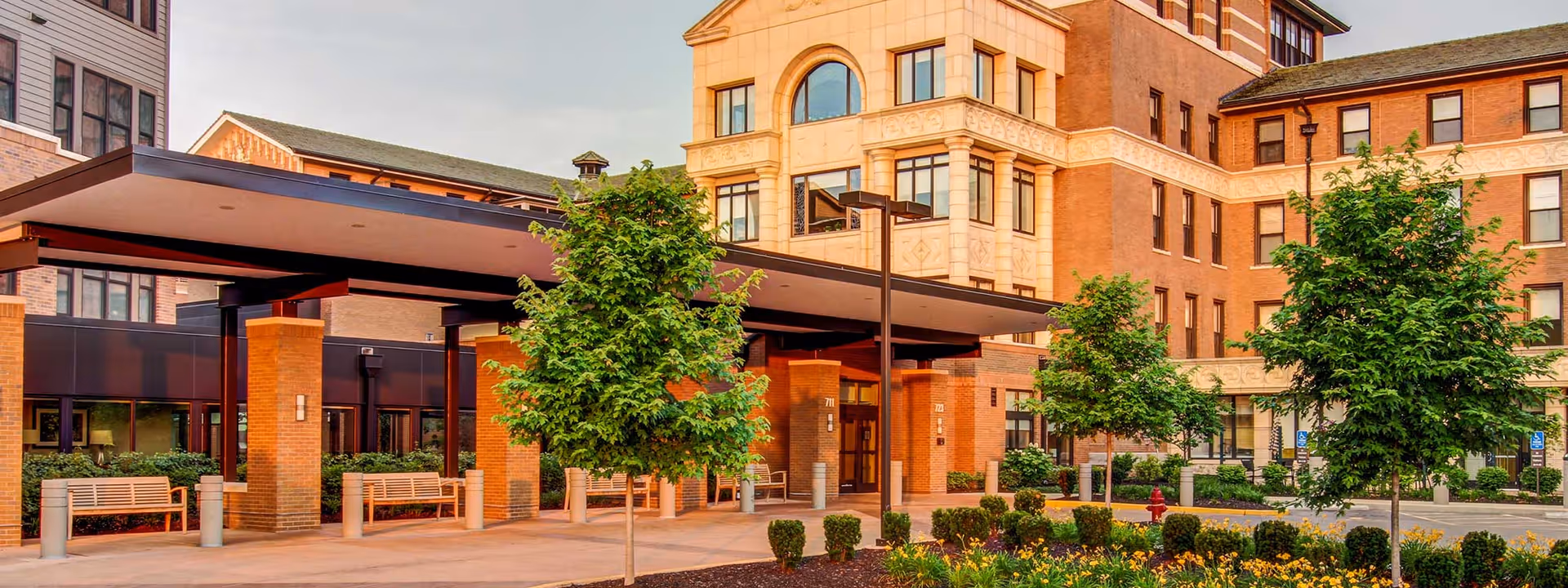 Entrance area of a senior living facility with a covered drop-off zone supported by brick pillars, surrounded by small trees, shrubs, and benches. The multi-story building in the background has large windows and a mix of brick and light-colored exterior walls.