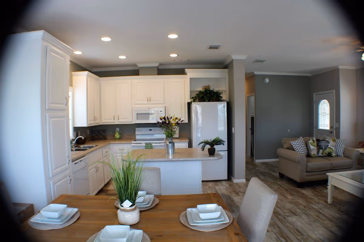 Interior view of a retirement community home showing a kitchen with white cabinets, a white refrigerator, stove, and microwave. In the foreground, there is a wooden dining table set with white dishes and a small plant centerpiece. To the right, a living area with a beige sofa adorned with patterned cushions is visible, along with a glass-top coffee table and a door with a decorative glass panel.