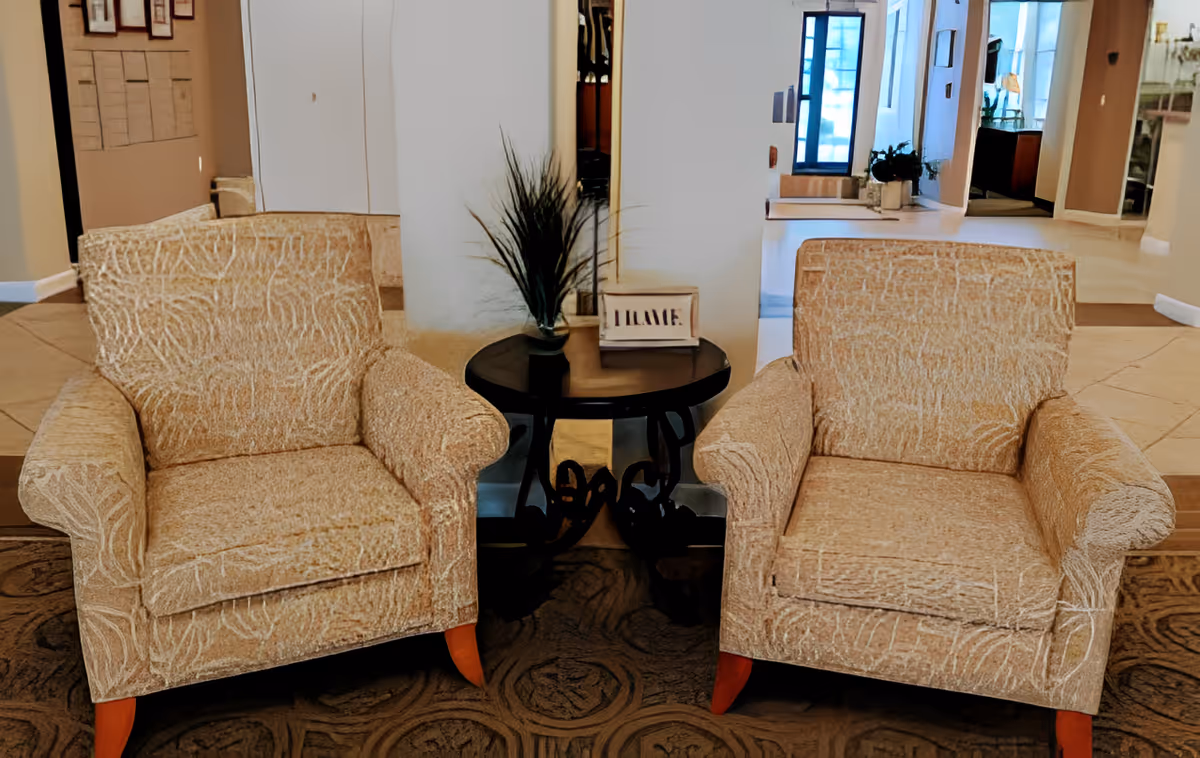 Two beige patterned armchairs with wooden legs are positioned on either side of a small round black table with decorative legs. On the table, there is a small plant and a framed sign that says 'WELCOME'. The background shows a hallway with light-colored walls and tiled flooring.