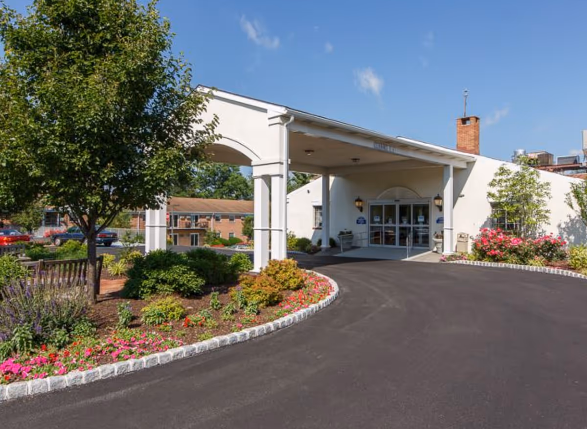 Entrance of Fort Washington Estates facility with a covered driveway, landscaped garden with flowers and bushes, a tree, and a clear blue sky.