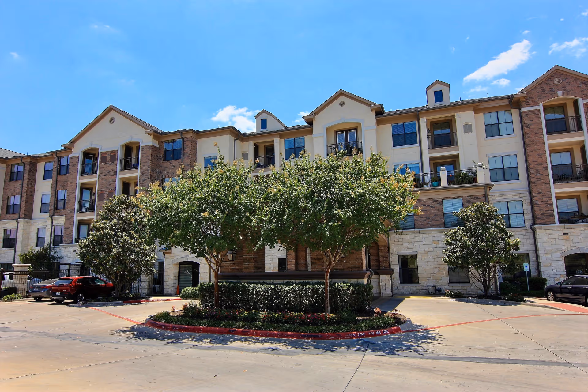 Exterior view of a multi-story senior living facility building with brick and light-colored stone facade, several windows, balconies, and a landscaped area with trees and shrubs in front. There are parked cars on either side of the entrance driveway under a clear blue sky.