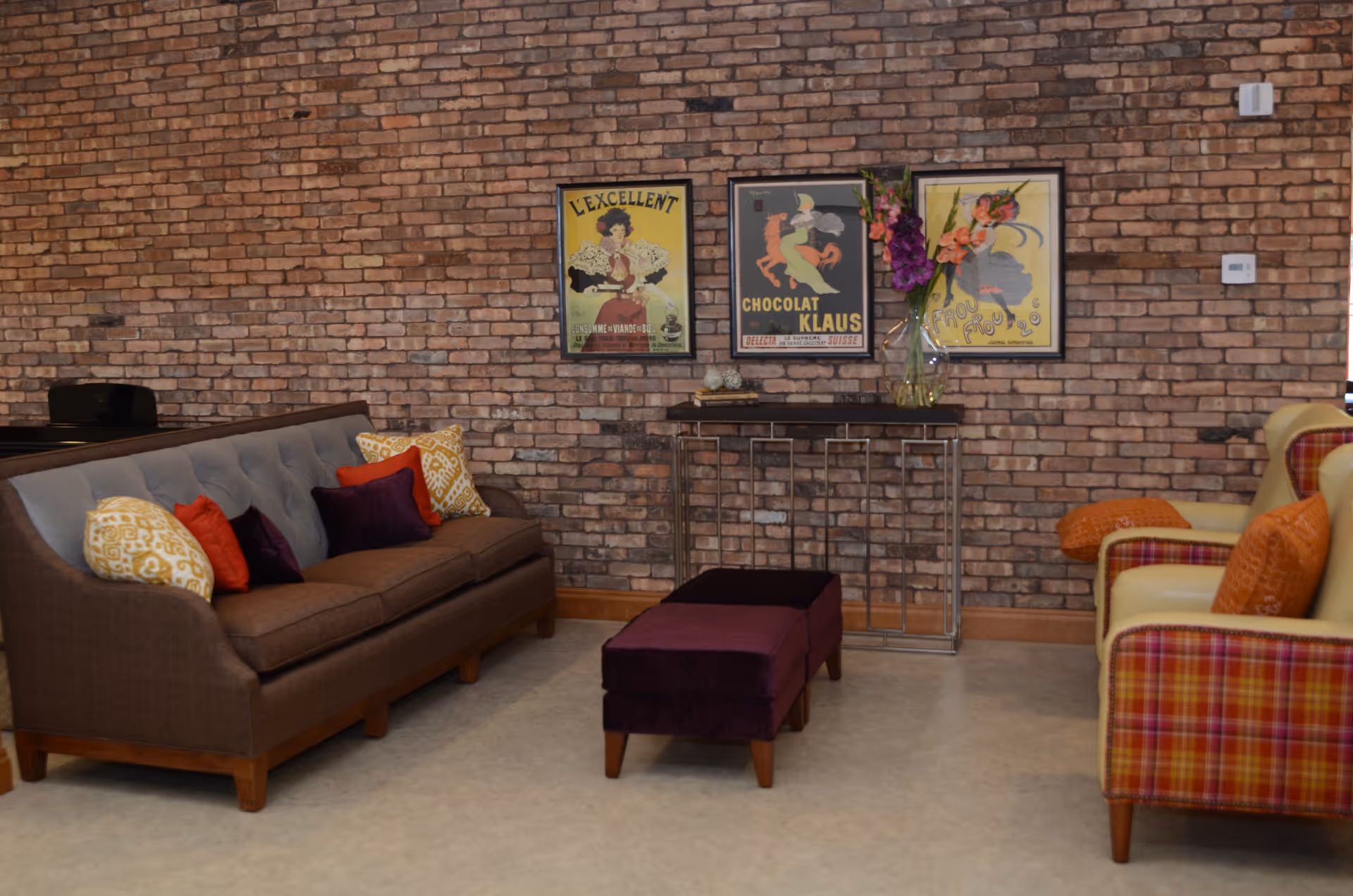 A cozy living room area with a brown sofa adorned with colorful pillows, two plaid armchairs with orange cushions, a small purple ottoman, and a console table against a brick wall. Three vintage-style framed posters and a vase with flowers are displayed on the wall above the console table.