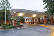 Entrance area of a senior living facility with a covered drop-off zone supported by brick columns, surrounded by trees and landscaped bushes.