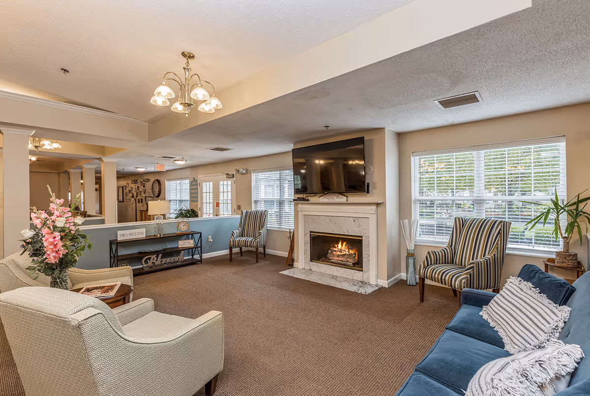 A cozy living room area in DeWolfe Place featuring a lit fireplace with a mounted flat-screen TV above it, a blue couch with decorative pillows, two striped armchairs, a beige armchair, a small round wooden table with a flower vase, and a console table with decorative items. Large windows with white blinds allow natural light to fill the room.