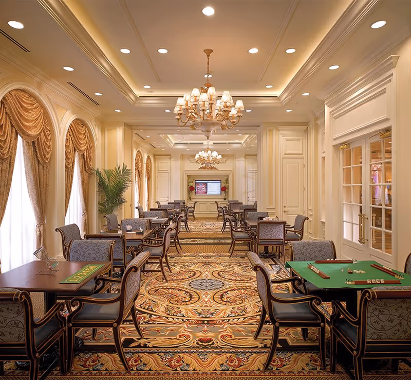Elegant game room with multiple tables and chairs arranged for playing games like mahjong, featuring ornate chandeliers, large windows with draped curtains, and a richly patterned carpet in a luxurious senior living facility.