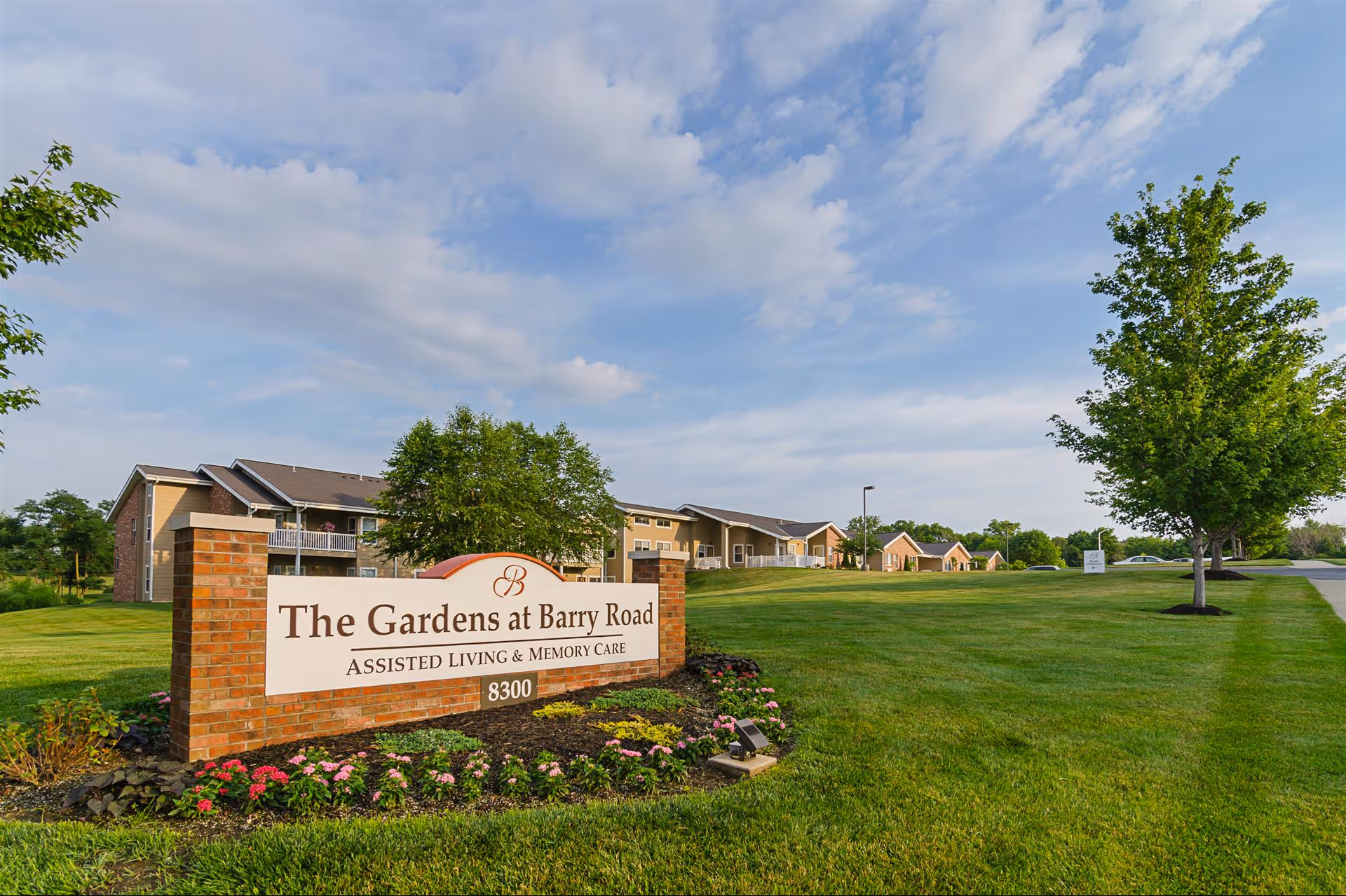 The entrance sign for The Gardens at Barry Road Assisted Living and Memory Care, surrounded by a well-maintained lawn, flowers, and trees, with the facility buildings visible in the background under a partly cloudy sky.