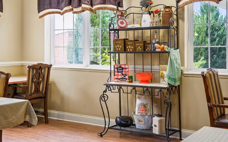 A cozy dining area with wooden chairs and tables covered with tablecloths. A decorative black metal shelf stands against the wall, holding various kitchen items including vintage-style canisters labeled flour, sugar, and coffee, a red bowl, a green checkered towel, and other small decorative objects. Large windows with brown valances let in natural light and show greenery outside.