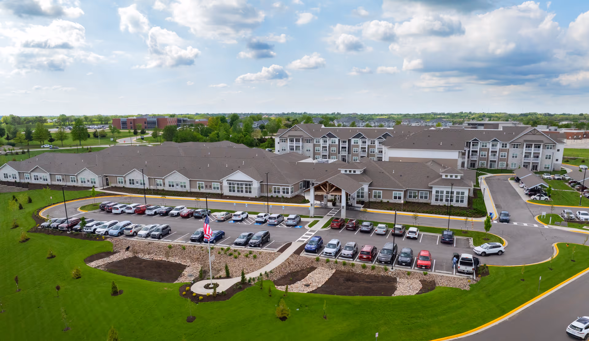 Aerial view of The Crestone Senior Living facility showing a large building complex with multiple wings, a parking lot filled with cars, landscaped green lawns, and an American flag on a flagpole in front. The sky is partly cloudy.