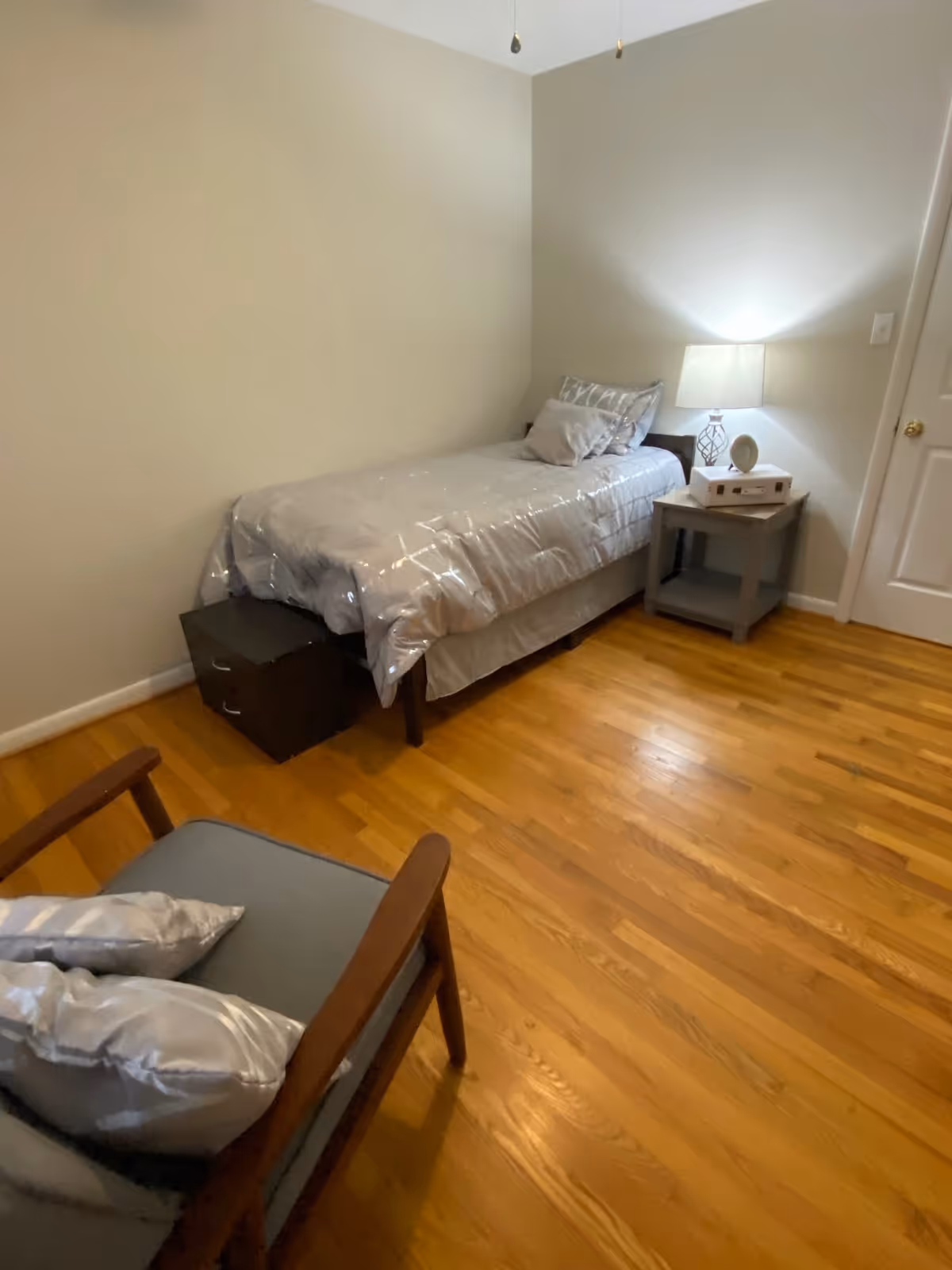 A small bedroom with a single bed covered in a gray comforter and pillows. Next to the bed is a gray nightstand with a lamp, a clock, and a white box on top. In the foreground, there is a wooden chair with gray cushions and pillows. The room has wooden flooring and beige walls.