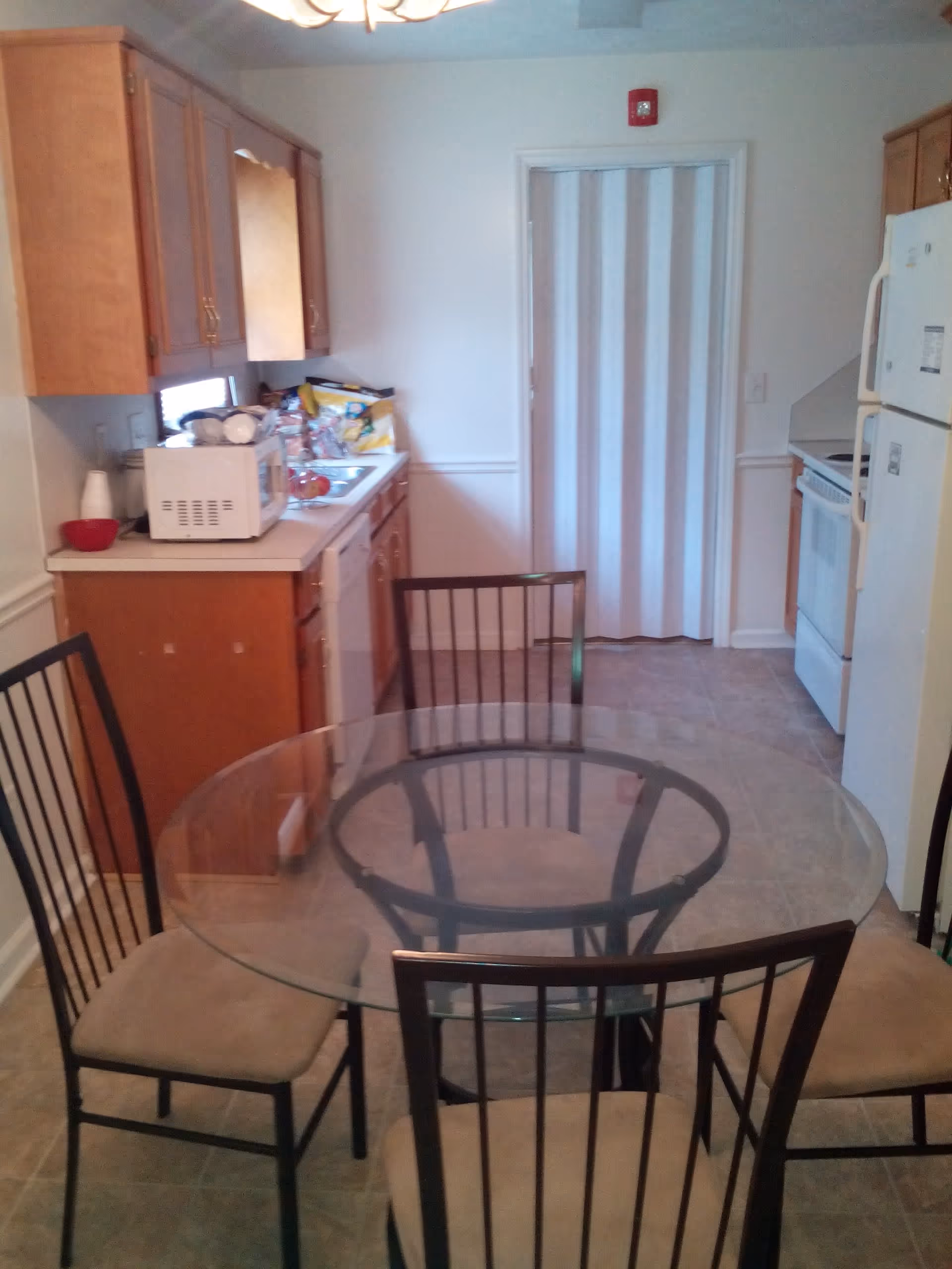 A kitchen and dining area with a round glass table surrounded by four chairs with beige cushions. The kitchen has wooden cabinets, a white refrigerator, a white stove, a microwave on the counter, and various kitchen items near the sink. A white folding door is visible at the back of the room.