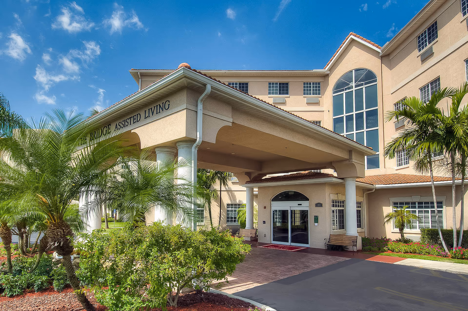Exterior view of The Bridge Assisted Living facility showing the entrance with a covered drop-off area supported by white columns, surrounded by palm trees and landscaped greenery under a blue sky with some clouds.