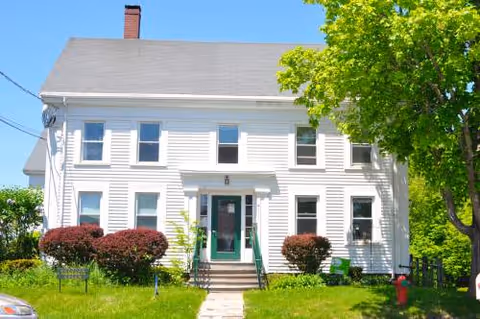 White two-story building with green door entrance surrounded by shrubs and trees.