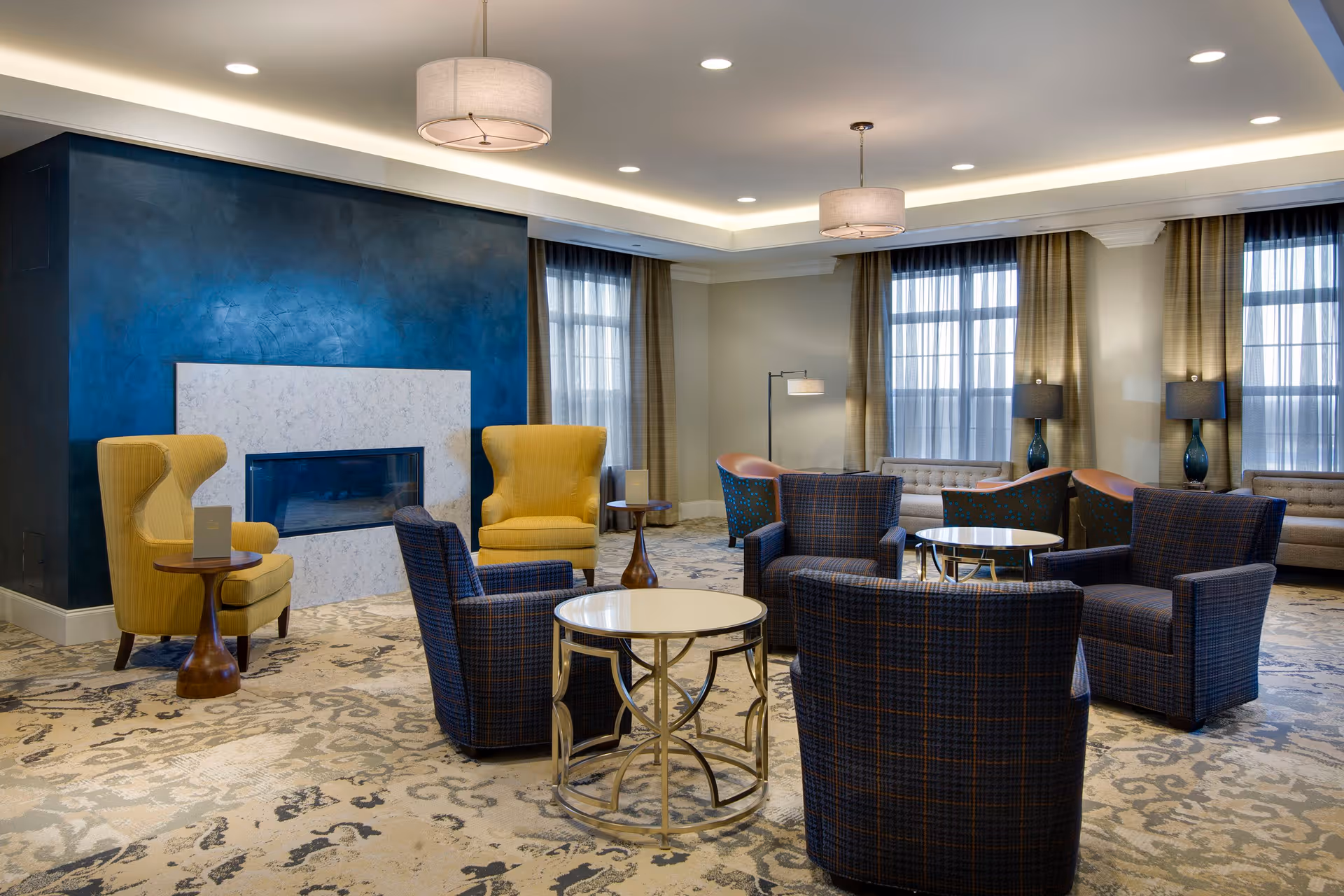 A spacious and well-lit living room area with multiple seating options including yellow armchairs, blue plaid armchairs, and beige sofas. The room features a modern fireplace with a dark blue accent wall, large windows with sheer curtains, and round tables with metallic bases. The ceiling has recessed lighting and two hanging light fixtures.